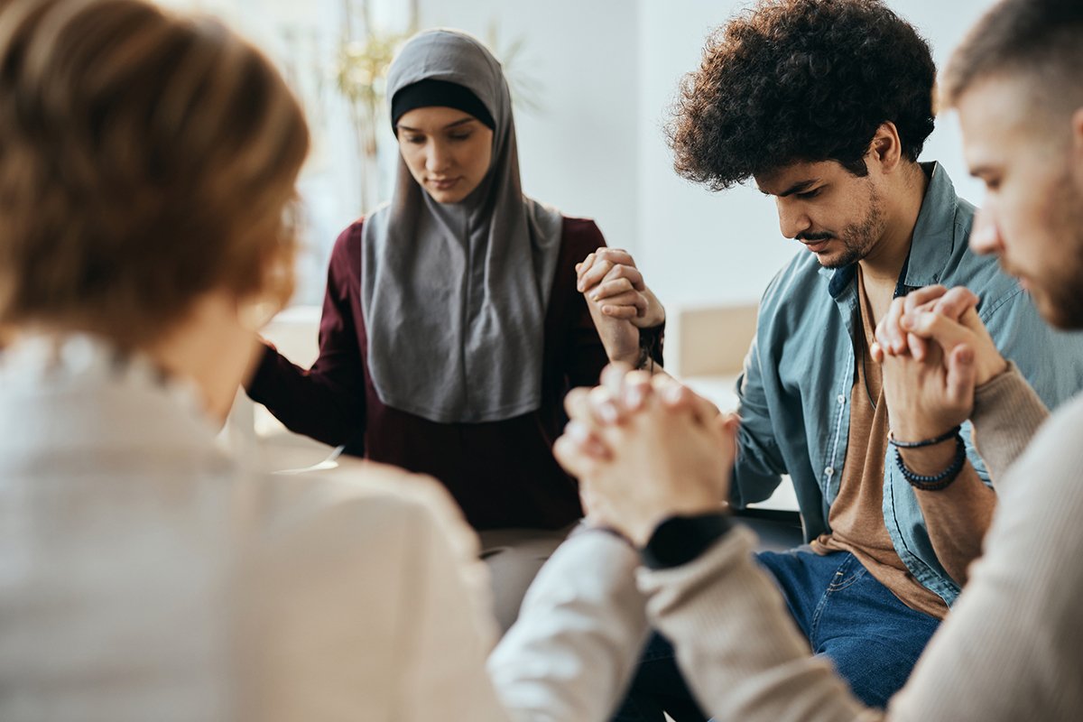Multiracial people holding hands during group therapy meeting. Focus is on Muslim man.